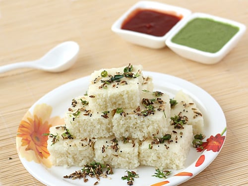 White coloured rava dhokla stacked on a plate served with green and red chutney