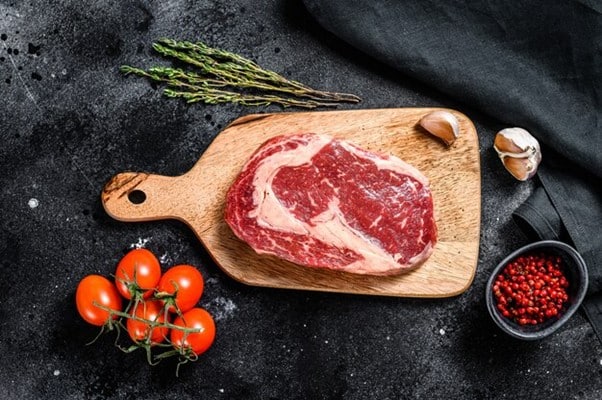 Raw ribeye steak on a wooden board with cherry tomatoes garlic peppercorns and fresh herbs