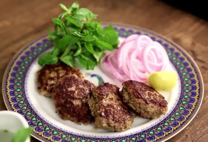 A plate of Galouti kebab with slices of onion a piece of lemon with mint leaves.